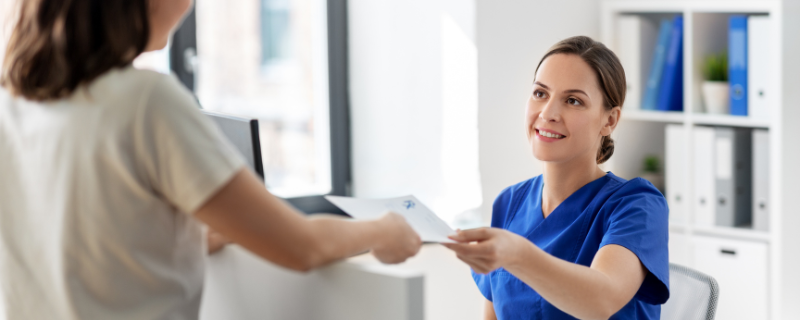 Imagem de uma mulher entregando um documento para outra, trajando uniforme azul em uma clínica.